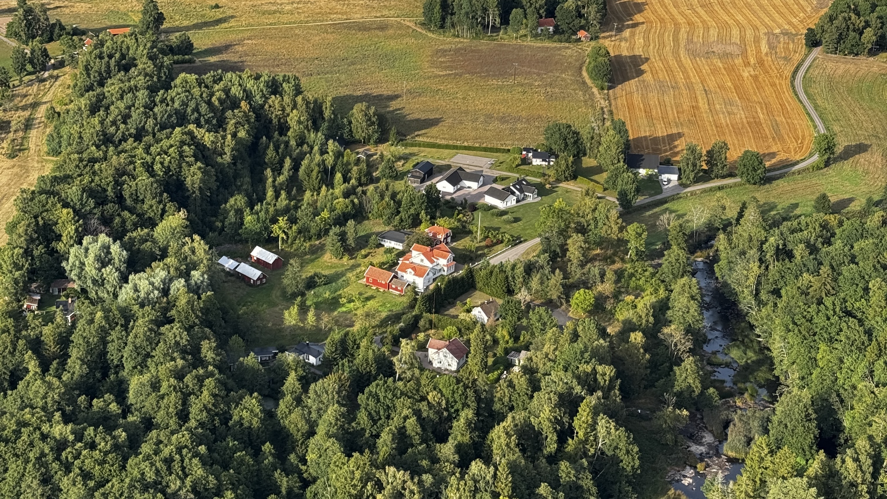 Flygbild i kvällssol: gårdens vita och röda hus bland lummiga träd, med ån Lidan som slingrar sig genom skogen och gula, skördade åkrar runt omkring.
