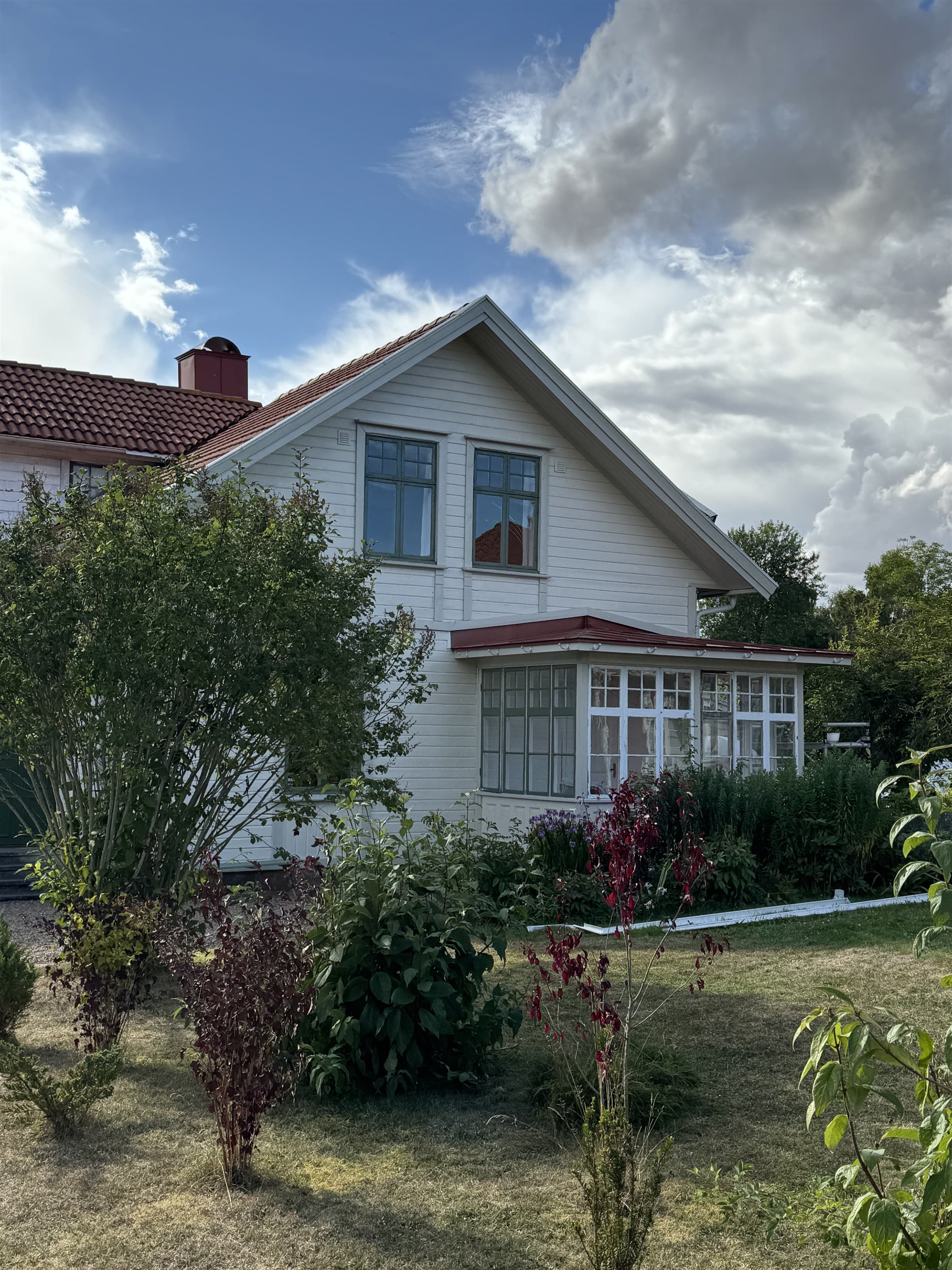 Huvudbyggnadens nymålade vita gavel med gröna fönsterbågar och en glasad veranda, bakom en trädgård med buskar under en blå himmel.