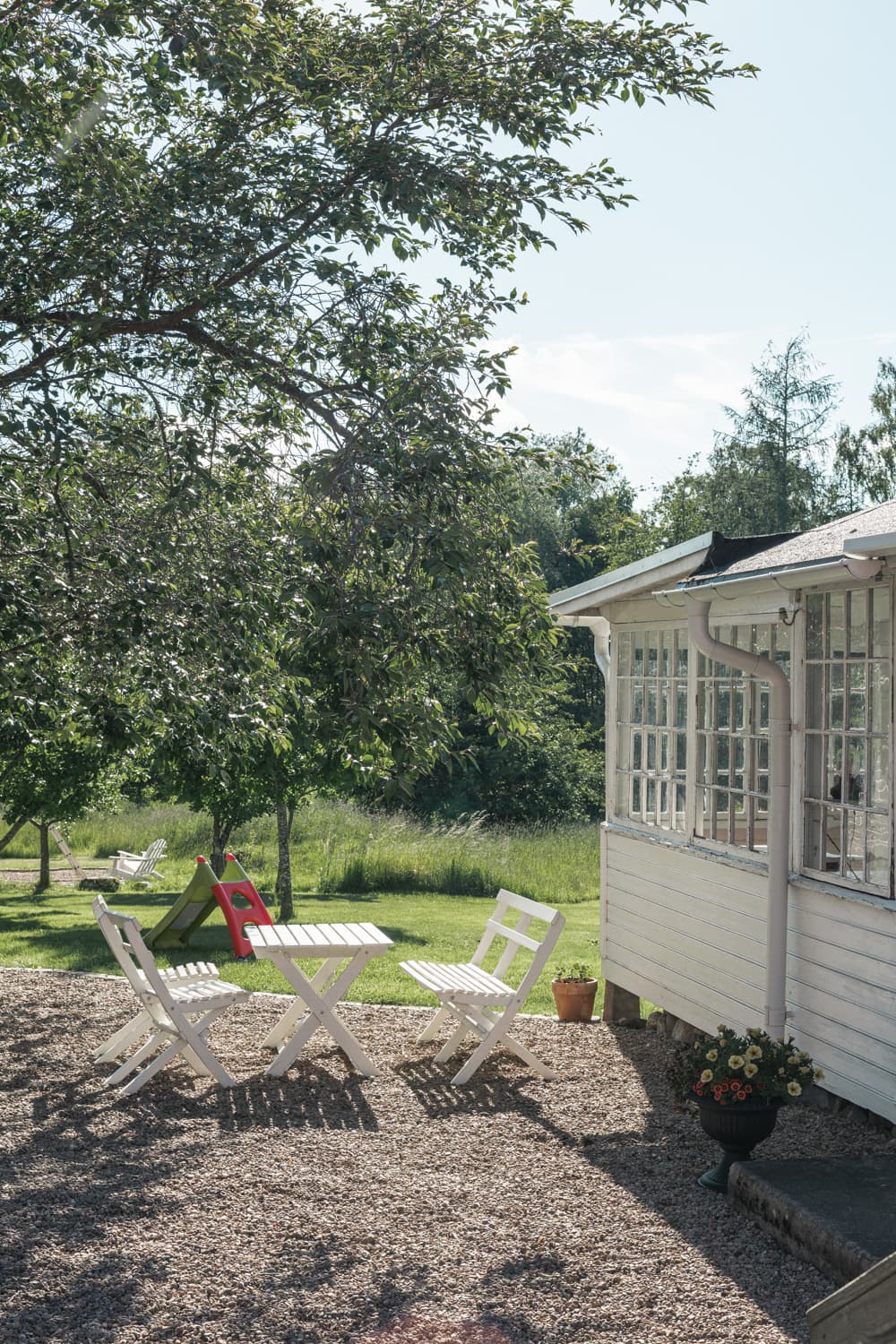 Paviljongens spröjsade fönster och vita panel mot trädgården, med ett bord på gruset under trädet.